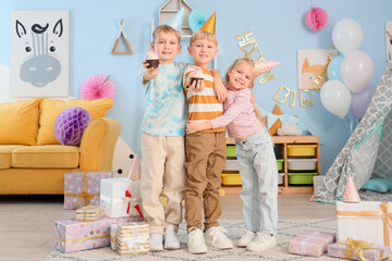 Cute little happy kids in party hats with cupcakes and gift boxes celebrating birthday at home