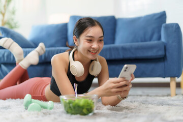 Happy asian fitness girl laying on floor using mobile phone app browsing internet, relaxing after exercising at home