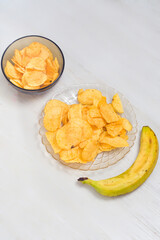Crispy potato chips served in a bowl and arranged on a clear plate alongside a ripe banana. The snacks are placed on a white wooden table, creating a simple yet appealing food scene.