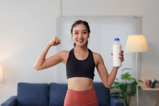 Smiling asian sportswoman flexing her biceps and holding a bottle of milk, promoting healthy lifestyle and calcium intake for strong bones