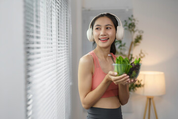 Young sporty woman enjoying healthy lifestyle eating a bowl of fresh salad while listening to music with headphones at home