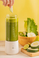 Generated imageHand placing a green apple slice into a portable blender with green detox smoothie, surrounded by cucumber, lime, and fresh lettuce.