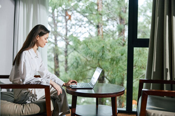 A focused young woman working on her laptop in a serene, green environment, wearing a casual white top and beige pants, captured in a bright, airy space