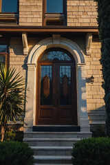 Fototapeta premium A tan brick building's entrance features a dark brown double door with arched transom and stone detailing. Steps lead up to the door, flanked by shrubs. Sunlight casts shadows on the façade.