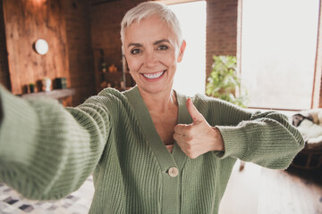 Charming elderly woman with short grey hair gives thumbs up and smiles warmly inside cozy living room