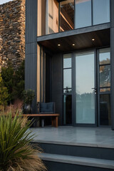 Modern home's exterior featuring dark gray siding, large glass doors, a stone wall, and a wooden bench on a paved patio with steps.  Landscaping includes grasses.
