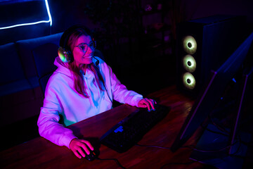 Young female gamer with headphones enjoying late night gaming session in neon-lit room, immersed in...