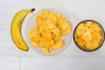 A single banana sits next to a plate and bowl of crispy, golden potato chips, arranged on a white wooden surface. A simple yet contrasting snack combination.