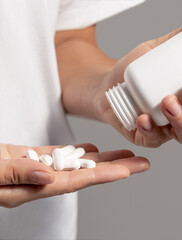 Woman pouring out white pills into hand from white plastic bottle closeup over white shirt