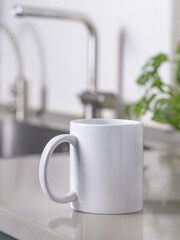 White coffee mug on grey kitchen table against kitchen interior with sink, closeup, mockup