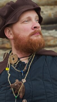Bearded man in medieval clothes stands near wooden house. Knight with key on neck and mouse in bosom looks into distance dreamy closeup horizon tilts