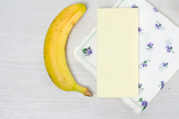 A ripe banana, a bar of white chocolate, and a delicate floral-patterned napkin arranged on a light...