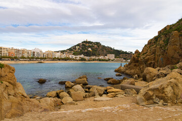 Obraz premium Vista desde Punta Sa Palomera hacia la bahía de Blanes, con rocas, mar y ciudad al fondo.