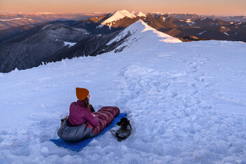 Young Woman in sleeping bag at winter mountains watching alpenglow sunrise. Bivouacs At Snowy...