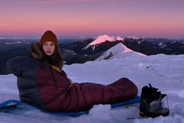 Young Woman in sleeping bag at winter mountains watching alpenglow sunrise. Bivouacs At Snowy...
