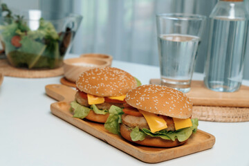 Focusing on double giant fried meat hamburger meal on square handle wooden board. Decorated by salad, homemade bun bread, beside drinking water on fast healthy food concept on white table. Infobahn.