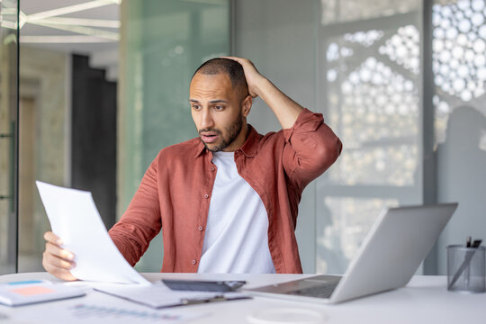 A man wearing a casual shirt looks surprised while reviewing documents in a stylish office, reflecting a work-related or unexpected discovery scenario.