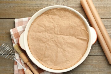 Ceramic casserole, baking parchment paper, brush and whisk on wooden table, flat lay