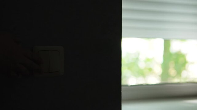Electric metal roller blinds on apartment window rise when control button is pressed. Person hand holds sewn on button for raising blinds and amount of light in room increases.