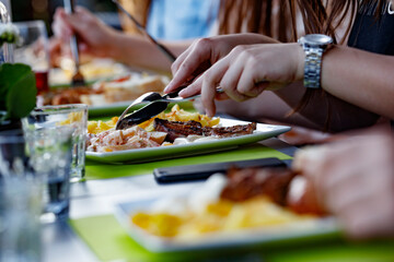 Group enjoying a meal outdoors.  Grilled meat, fries, and a salad are on the plate.  Hands are actively eating.