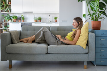 Happy woman relaxed lying on sofa with laptop on laps looking at screen watching interesting video or movie. Pleased female resting with portable computer enjoy internet time on couch at cozy home.