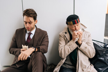 Sad businessman in suit and hungry senior homeless man sitting on piece of cardboard on city...