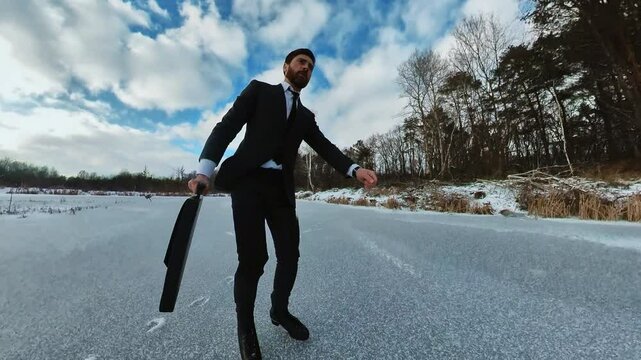 Footage of ice skating young Caucasian man with beard holding suitcase in hand. Shot of good-looking businessman in black suit on way to work. Outdoors. Lake, out of town