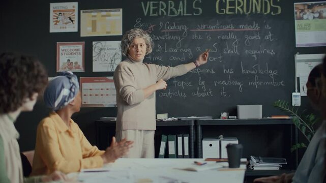 Middle-aged female teacher standing in classroom, pointing at text on blackboard, explaining gerund form of verb to group of diverse students during English lesson