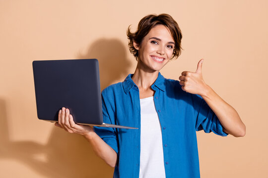 Portrait of successful young woman python web developer in blue shirt thumb up using netbook for projects isolated on beige color background