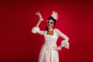 A young woman poses in a baroque costume against a vibrant red background, blending vintage style with modern flair and playful accessories