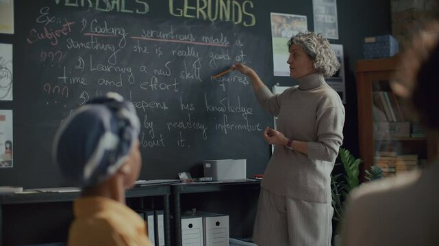 Female teacher pointing at blackboard, explaining English grammar lesson on gerunds to her students in language school