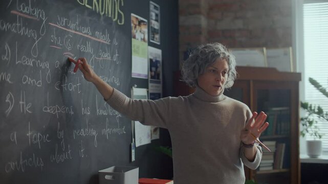 Mature female teacher with grey hair pointing at sentence written on blackboard and explaining English grammar rules during lesson in classroom