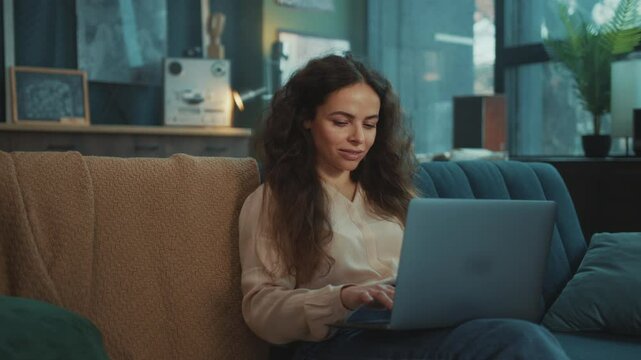 Portrait of smiling attractive focused young woman with beautiful hair in beige blouse typing on keyboard of laptop while sitting on sofa in modern living room. Indoors