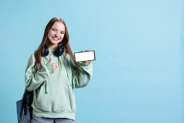 Cheerful teenager holding mockup smartphone, doing thumbs up sign gesturing, recommending it. Jolly...