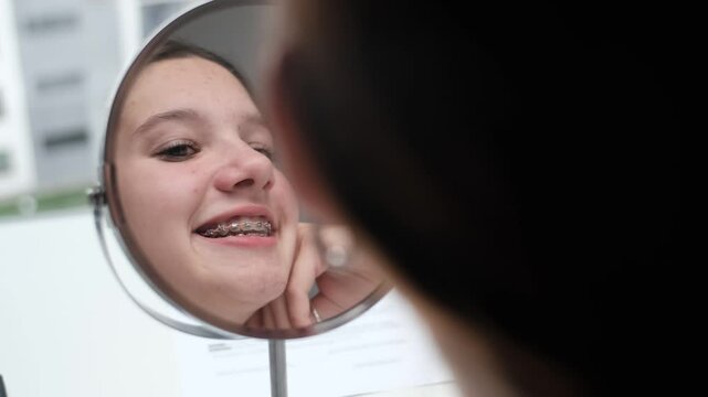 Teenage girl looks in mirror admiring her smile in braces. The concept of self-acceptance in braces. 