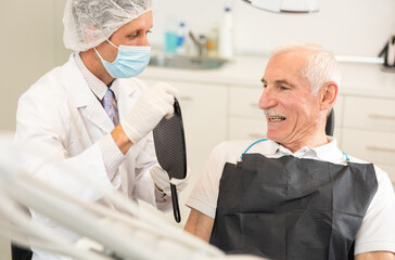 During appointment, dentist demonstrates to patient result of cosmetic dental filling. Man dentist specialist holds mirror in front of senior man patient and suggests evaluating effect of treatment.