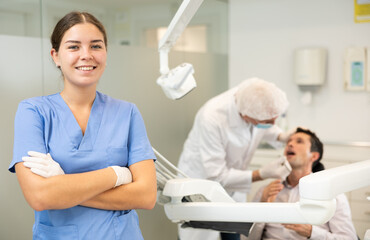 Obraz premium Young woman nurse in blue suit uniform is standing in dental office, clinic employee in suit uniform near dentists workplace. Doctor enjoys working in profession.