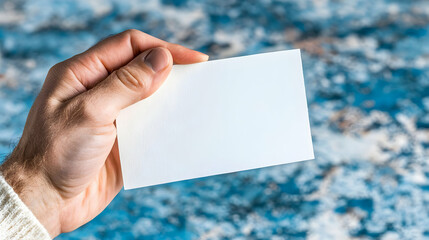 Close-up of a male hand holding a blank white card against a blurred blue and white background.