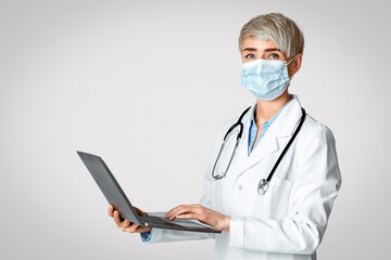 Visit at therapist concept. Woman doctor in protective mask holds laptop in her hands and writes medical history, isolated on light background, studio shot