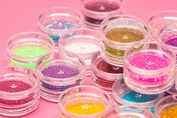 Collection of small plastic containers filled with various colors of glitter, on a pink surface. The vibrant and diverse colors of the glitter. Crafts, makeup, decoration concepts