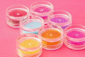 Collection of small plastic containers filled with various colors of glitter, on a pink surface. The vibrant and diverse colors of the glitter. Crafts, makeup, decoration concepts