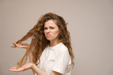 A frustrated, dissatisfied young woman touching ends of her hair and is horrified by the dry, damage and unruly hair and split ends of dry hair. 
