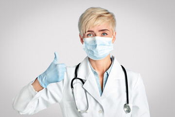 Great medical care, prevent disease and quarantine. Middle aged woman doctor in protective mask, white coat, with stethoscope, in rubber gloves shows thumbs up isolated on blue background, studio shot