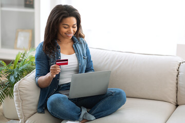 Naklejka premium A young woman sits cross-legged on a comfortable sofa, smiling while using her laptop. She holds a credit card, engaged in an online shopping activity in a bright living space, copy space