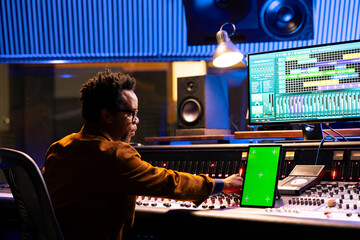 Sound technician mixing tracks next to greenscreen tablet in control room, operating panel board...