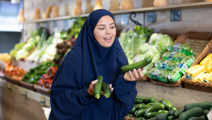 Pleased young woman customer taking fresh cucumbers at the counter in large greengrocery