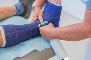 Nurse in clinic cutting strong fiberglass cast on patient's leg with plaster saw taking precautions, patient sitting quietly, professional help concept,