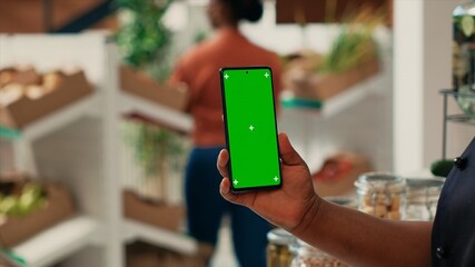 Vendor showing phone with greenscreen layout at supermarket, holding blank copyspace mockup on display. African american seller working at local grocery store, isolated chromakey screen.