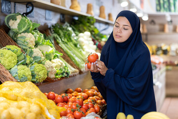 Attentive young Muslim woman purchaser choosing tomatoes at the counter in large grocery store