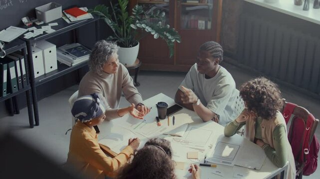 Mature female teacher and diverse students having group discussion during English class, sitting around table scattered with papers, notebooks, pens and coffee cup. View from above